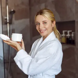 A woman in white robe holding up a jar of cream.