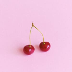 Two cherries are sitting on a pink surface.