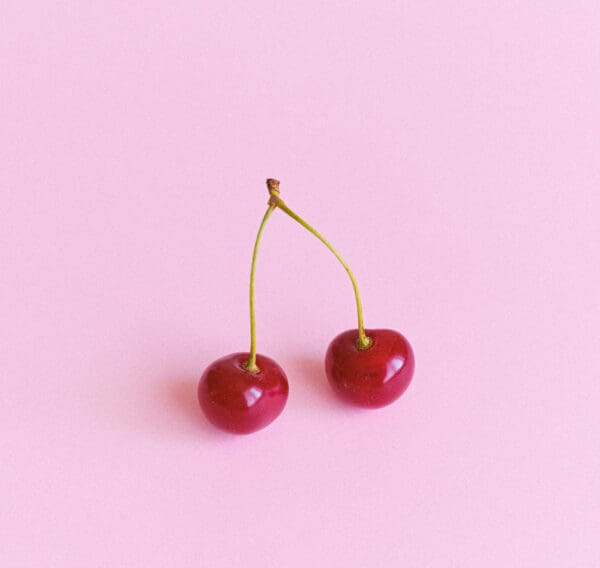 Two cherries are sitting on a pink surface.