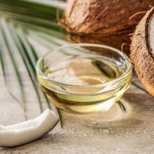 A bowl of oil next to coconut and palm leaves.