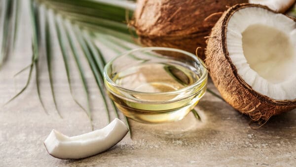 A bowl of oil next to coconut and palm leaves.