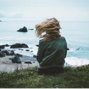 A woman sitting on the grass looking out at the ocean.