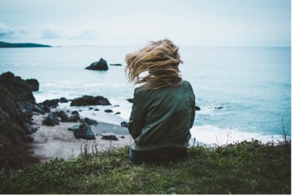 A woman sitting on the grass looking out at the ocean.